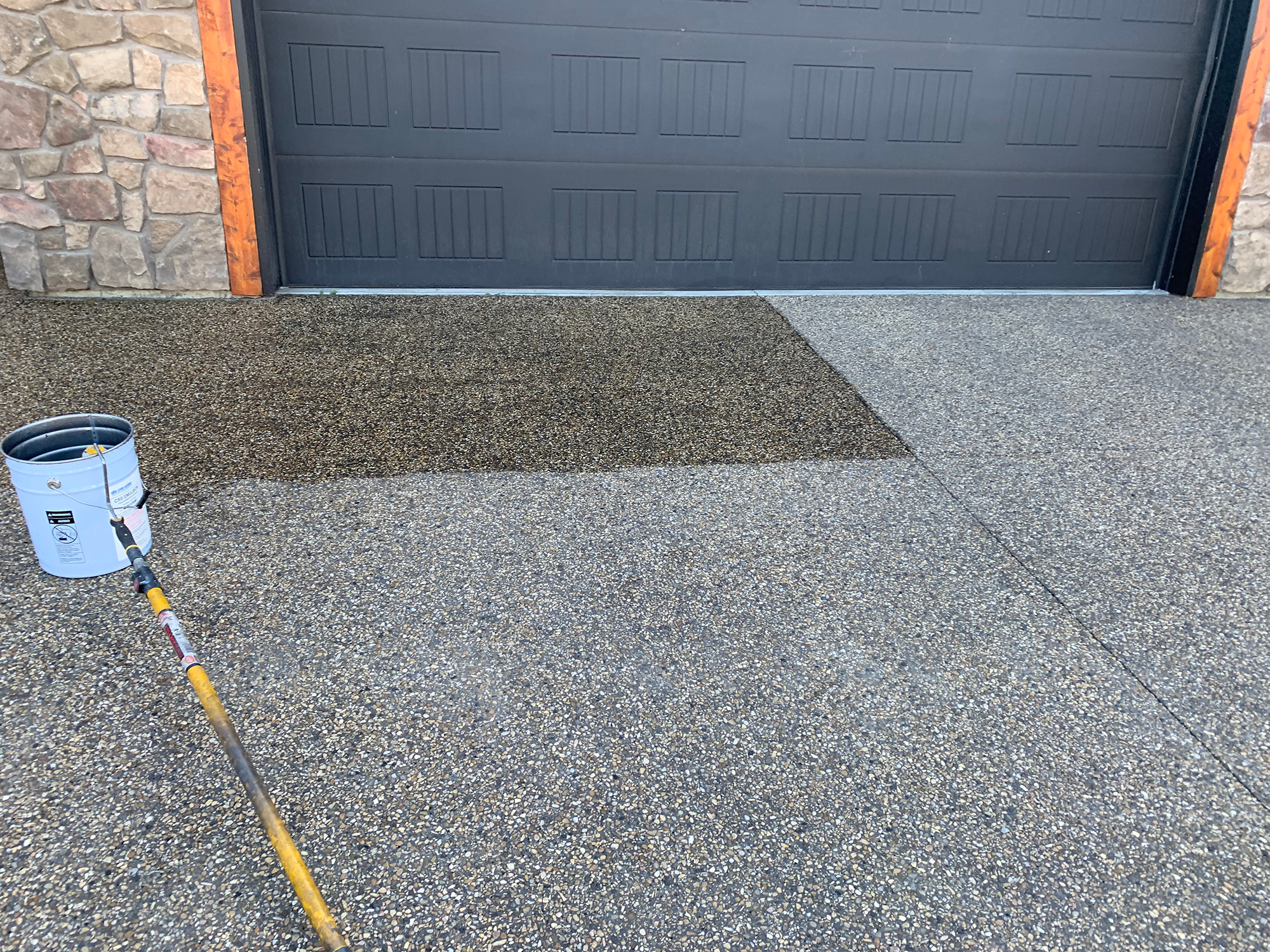 A textured concrete driveway with a section coated with sealant, a paint bucket, and a pole, in front of a black garage door with stone and wood accents.