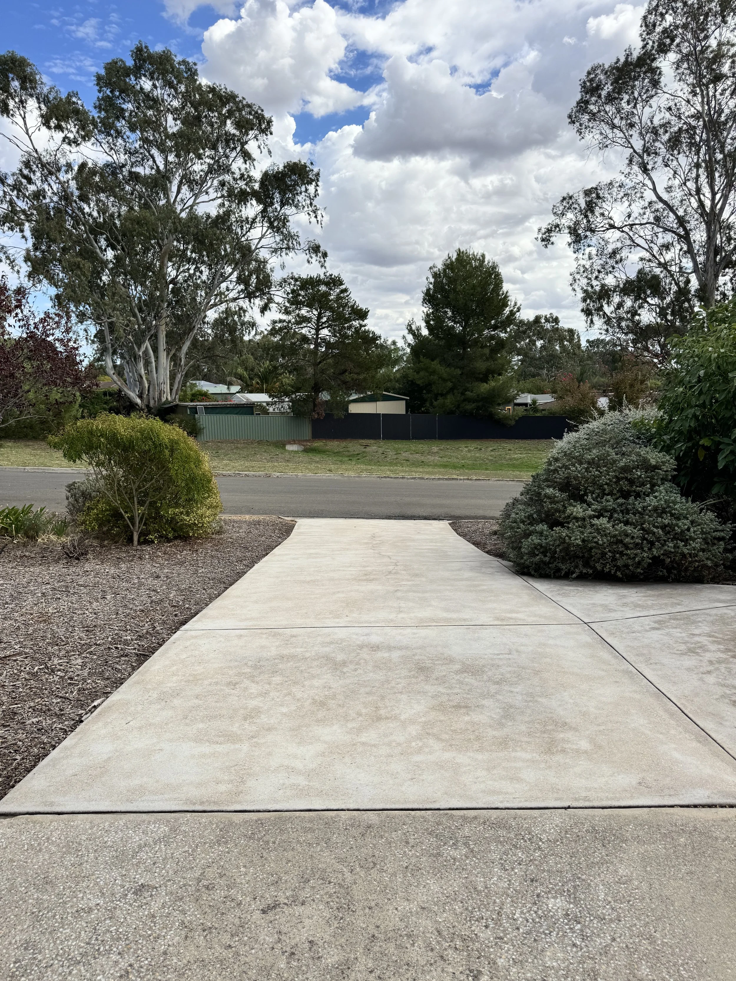 Concrete sidewalk leading to a street, with bushes on both sides, trees and a fence in the background under a partly cloudy sky.