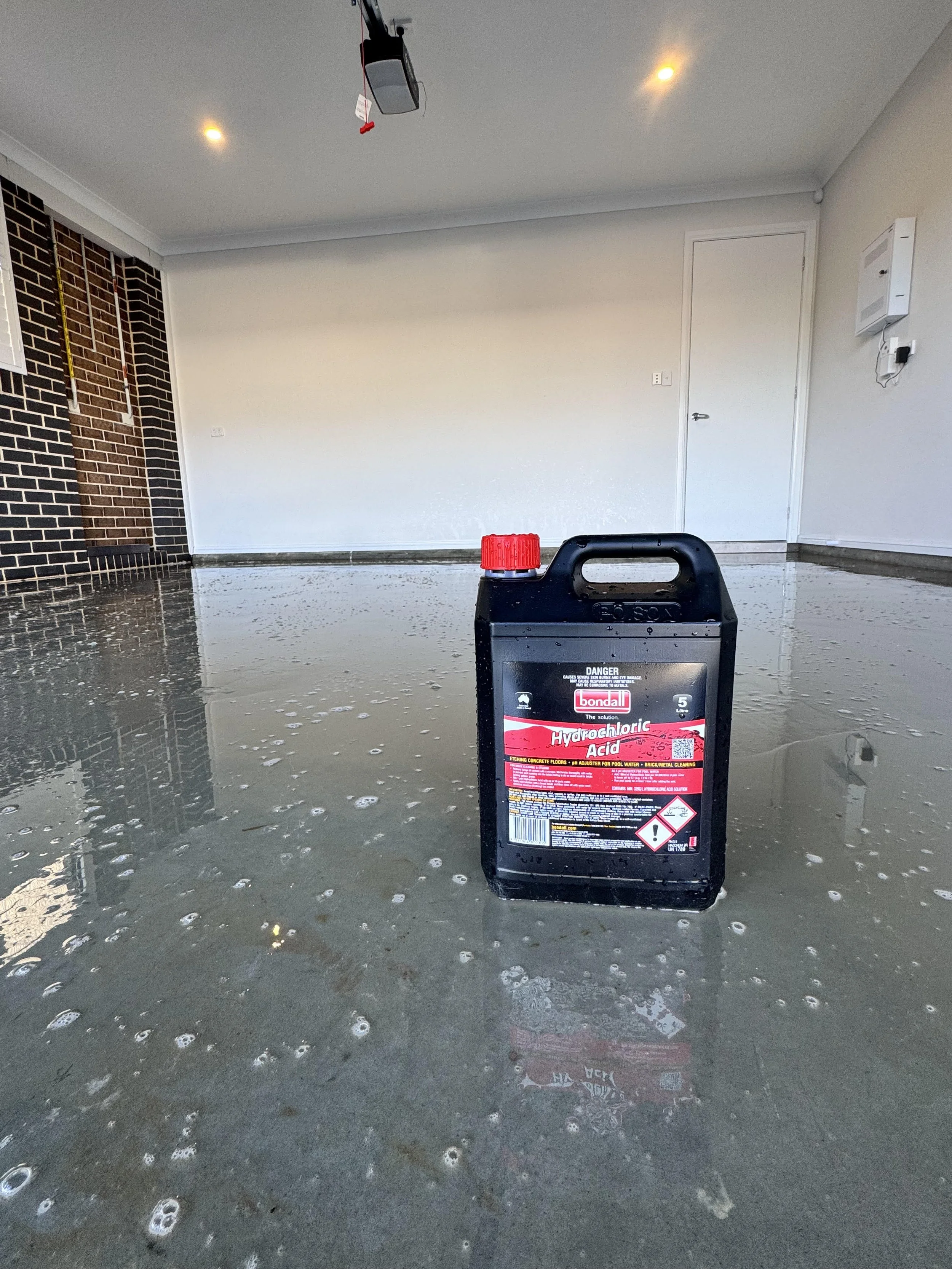 A container of hydrochloric acid placed on a wet, shiny floor in a garage or workshop with a brick wall and closed door in the background.