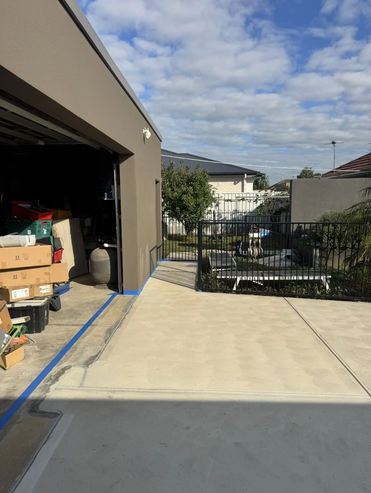 View of a backyard with a newly poured concrete patio, a black metal fence, a small tree, and neighboring houses under a partly cloudy sky. There are boxes and tools near a garage opening on the left.
