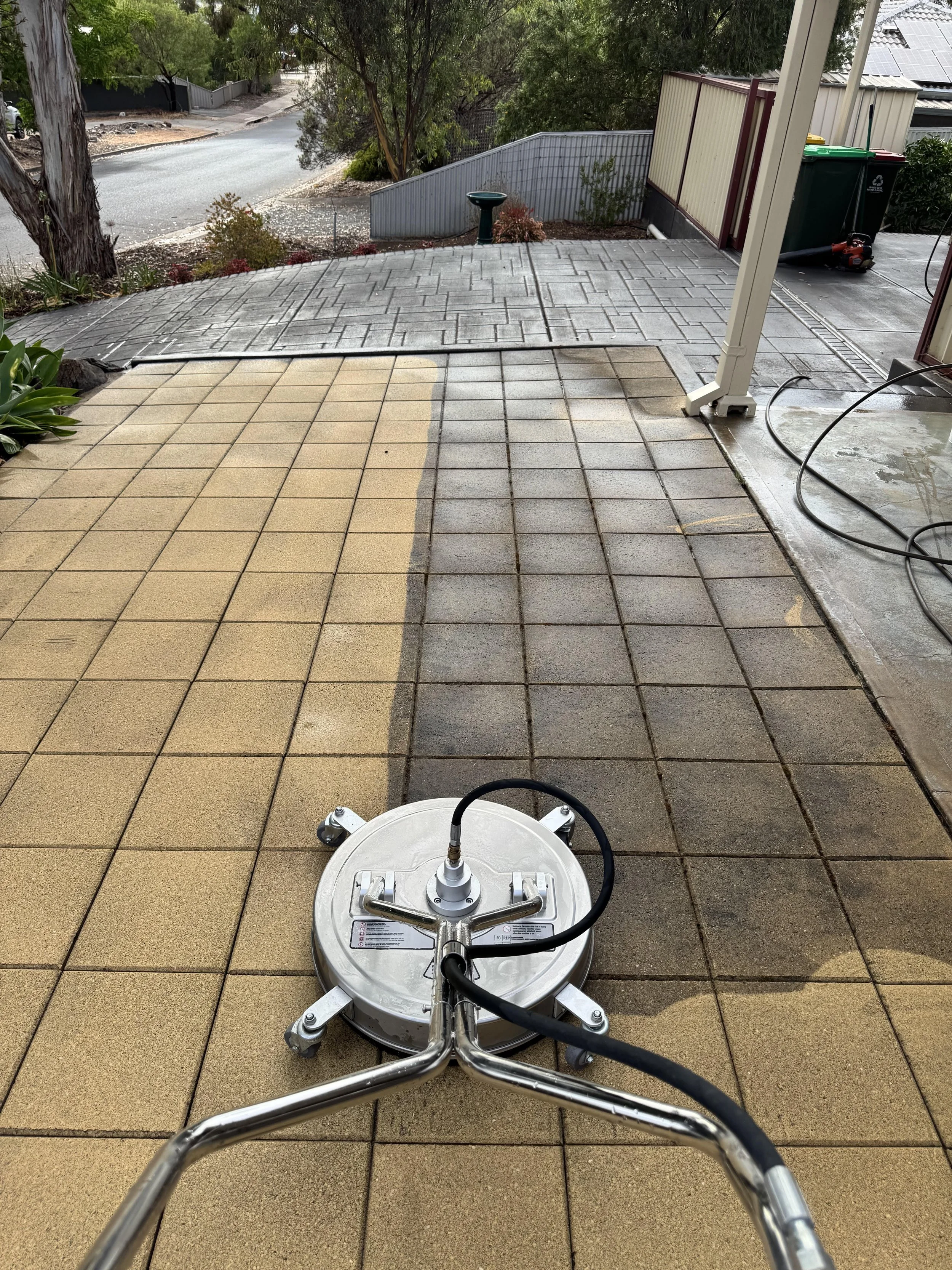 A pressure washing machine cleaning outdoor tiled patio with beige and gray tiles on a residential property.
