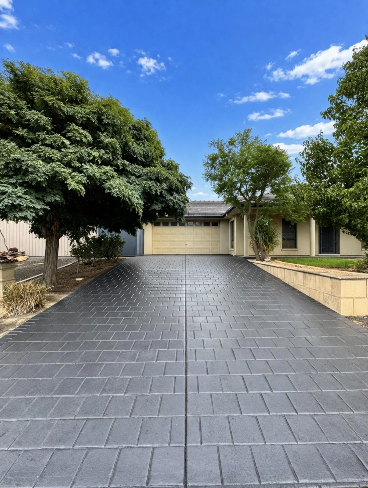 View of a house with a driveway made of dark interlocking pavers, trees on both sides, and a bright blue sky with scattered clouds.