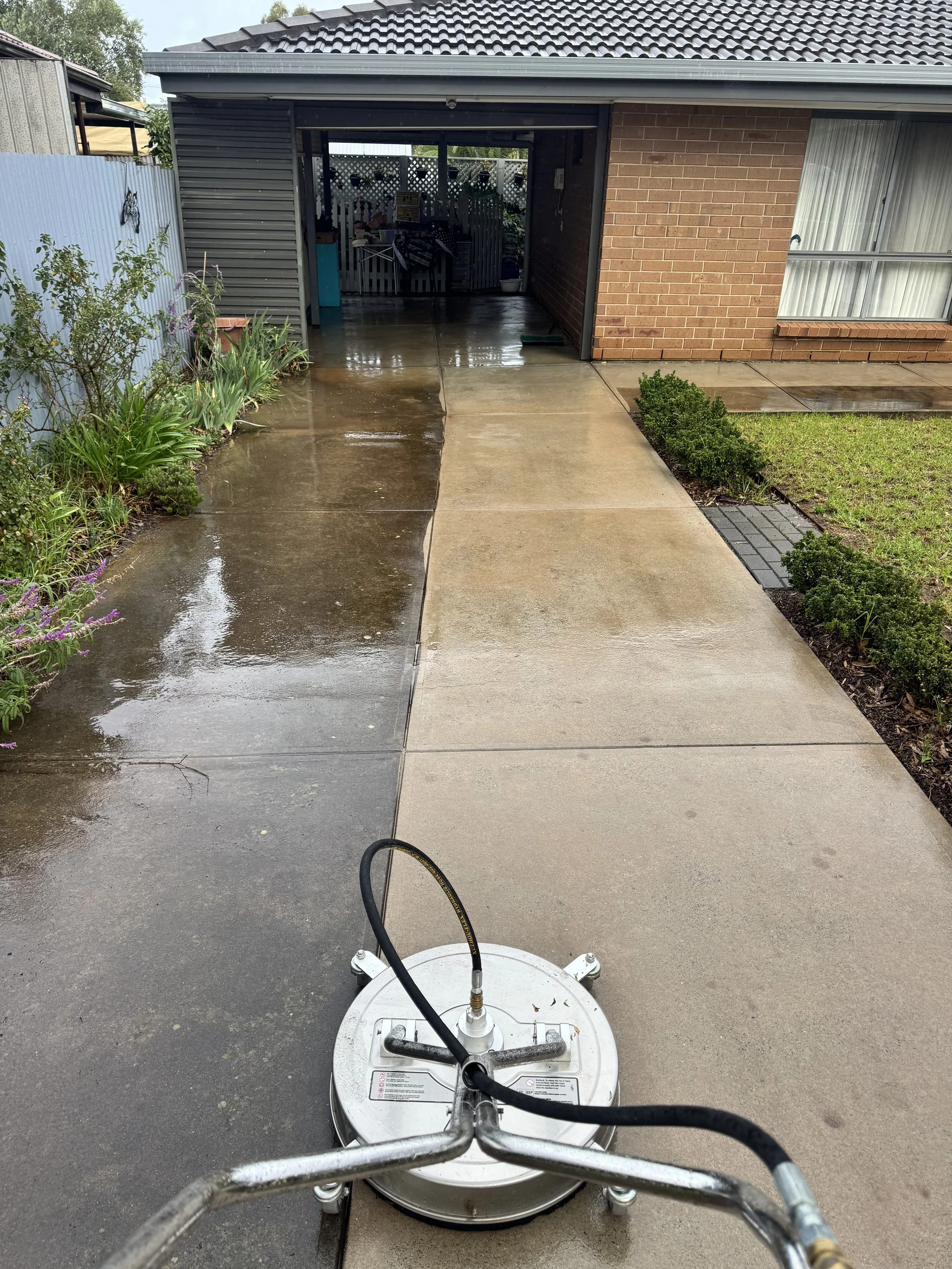 View of a pressure washer on a concrete driveway, with a wet section indicating recent cleaning, in front of a house with a brick wall, window, and a garage. There are garden plants on the left and a small bush on the right.