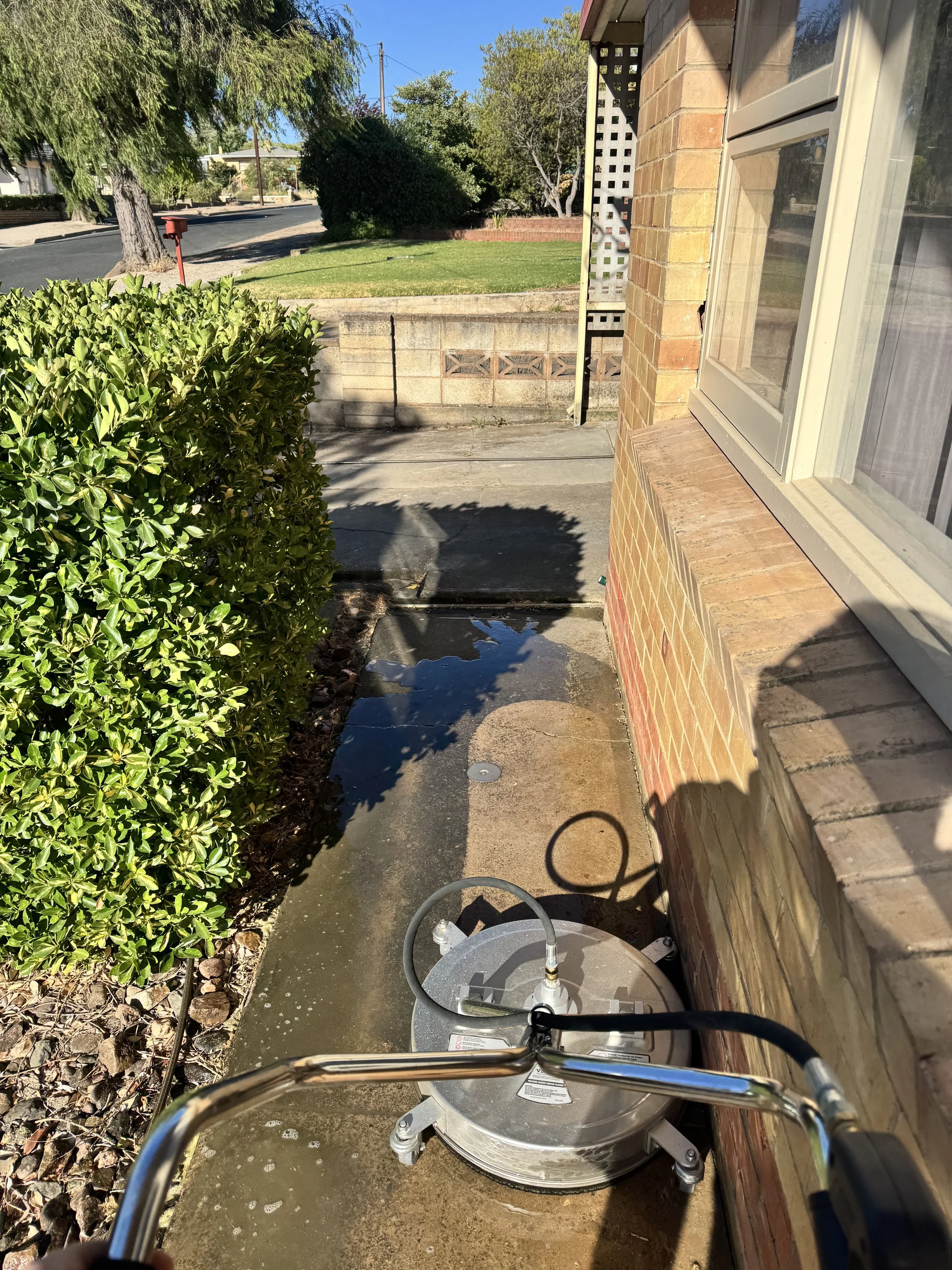The image shows a concrete sidewalk on the side of a house with a metal vaporizer machine placed on the ground. To the left, there's a green shrub, and in the background, a street with trees and a mailbox. The house has a brick exterior, and a window is visible on the right side of the image.