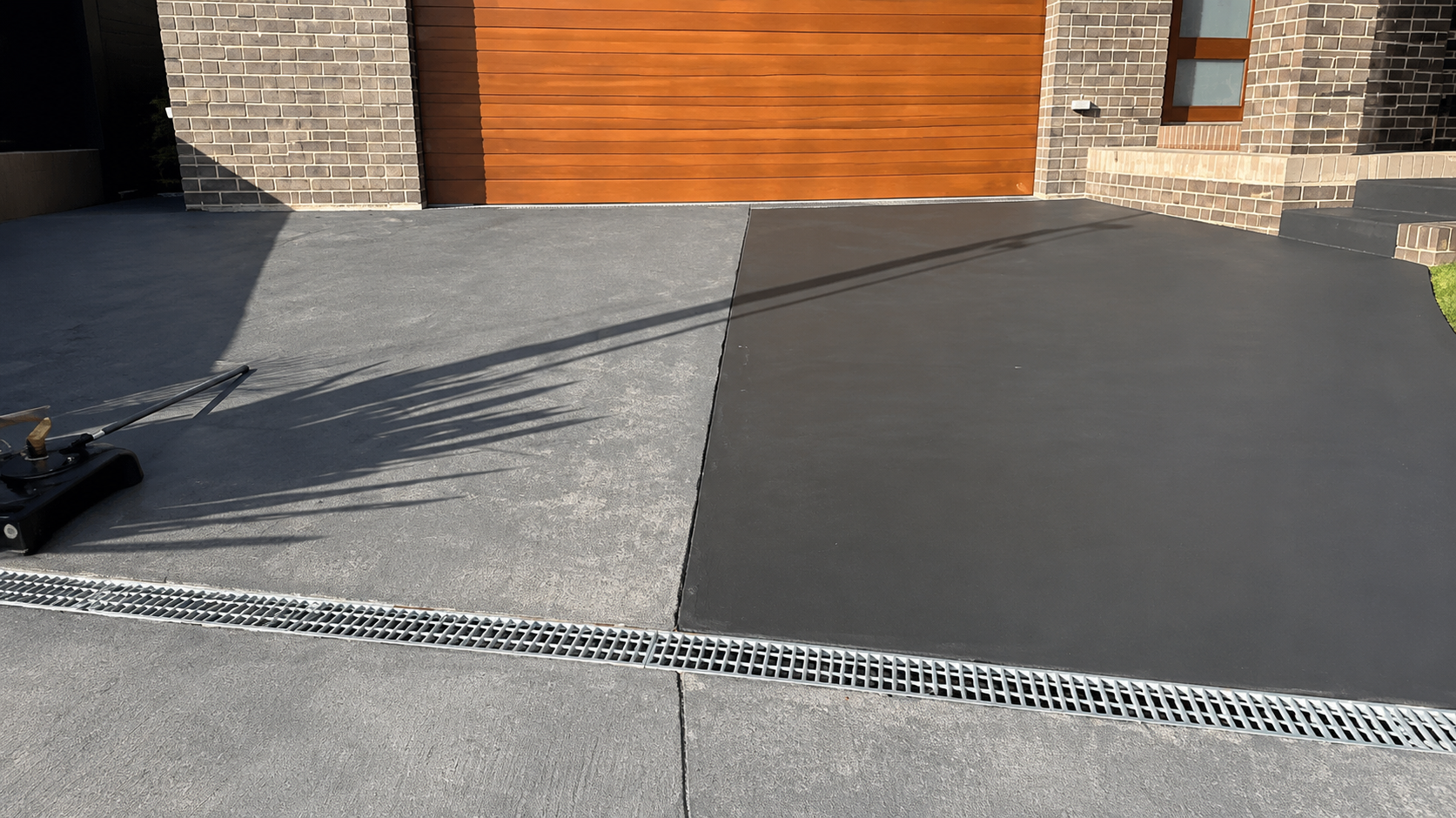 A driveway with a section of light gray concrete and a section of dark gray asphalt in front of a modern house with brick and wooden exterior walls.