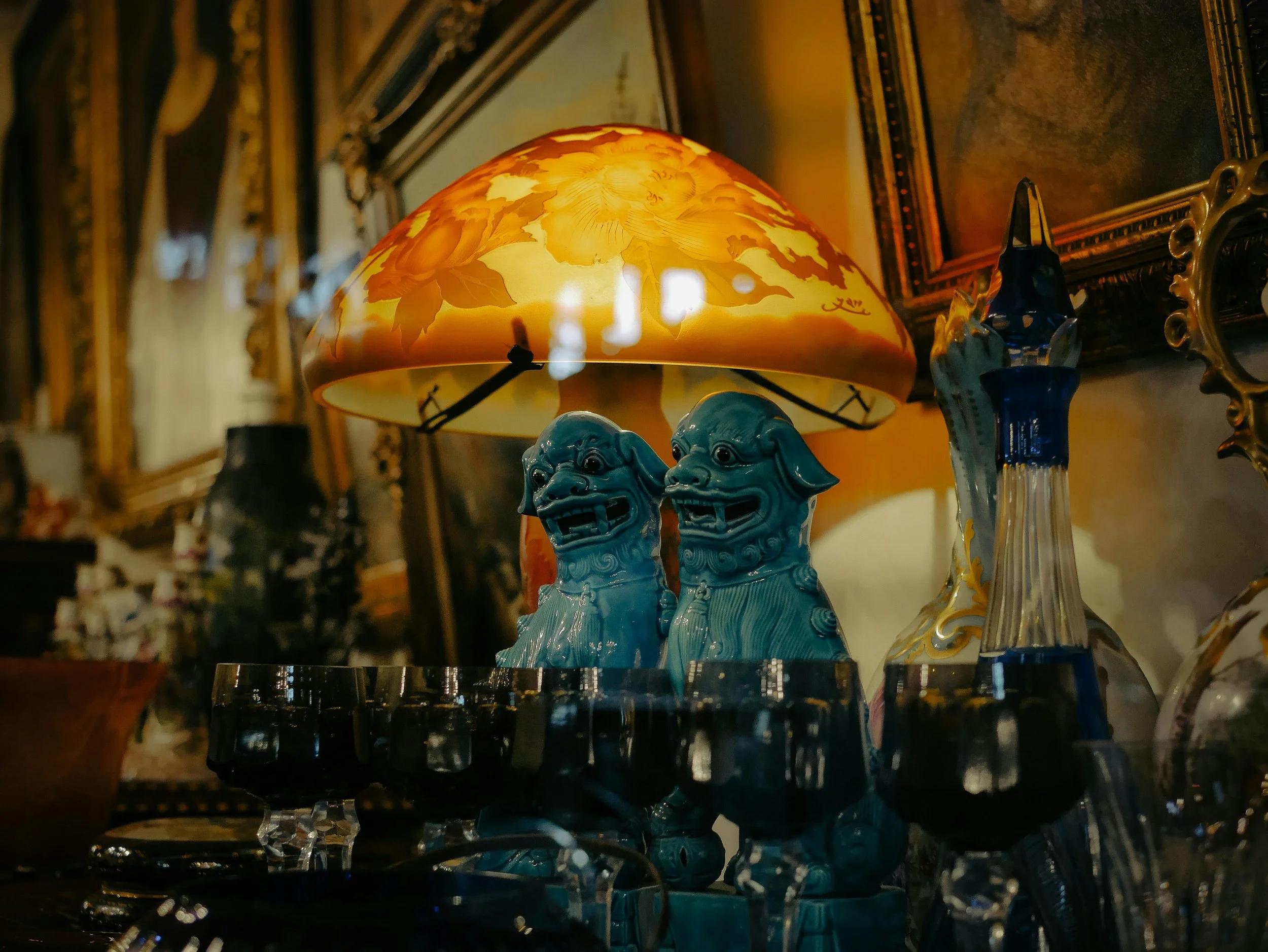 Decorative table lamp with floral shade, set on a glass table with blue ceramic figurines, amid framed paintings in a dimly lit room.