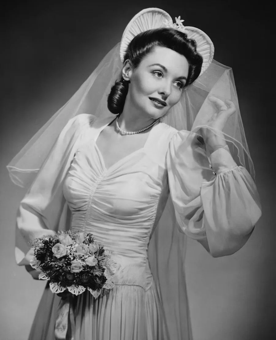 Black and white portrait of a woman in a vintage wedding dress with a veil, holding a bouquet of flowers, and wearing a pearl necklace.