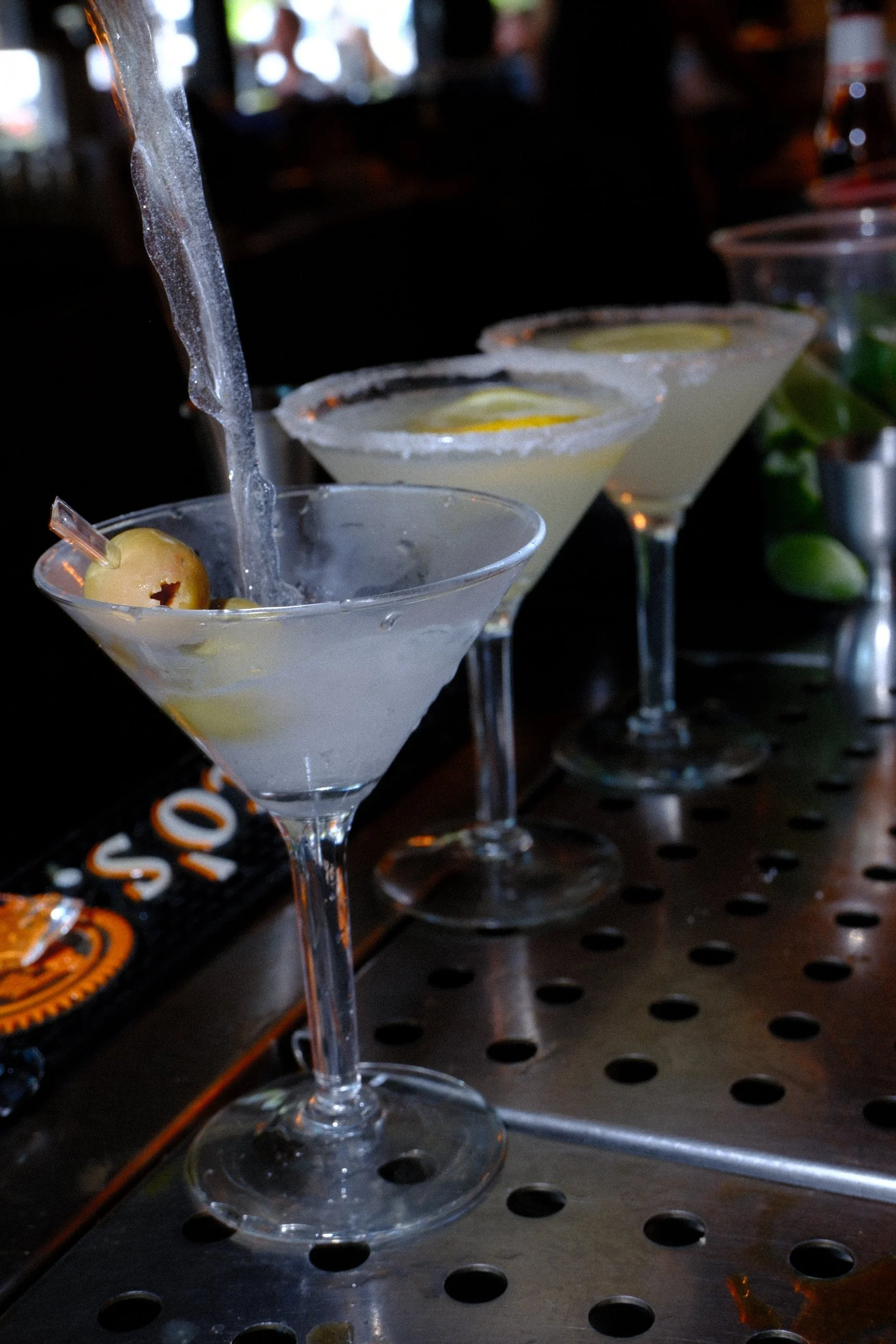 Frosted martini glass with a cocktail and olive garnish on a bar, with additional martinis in the background.