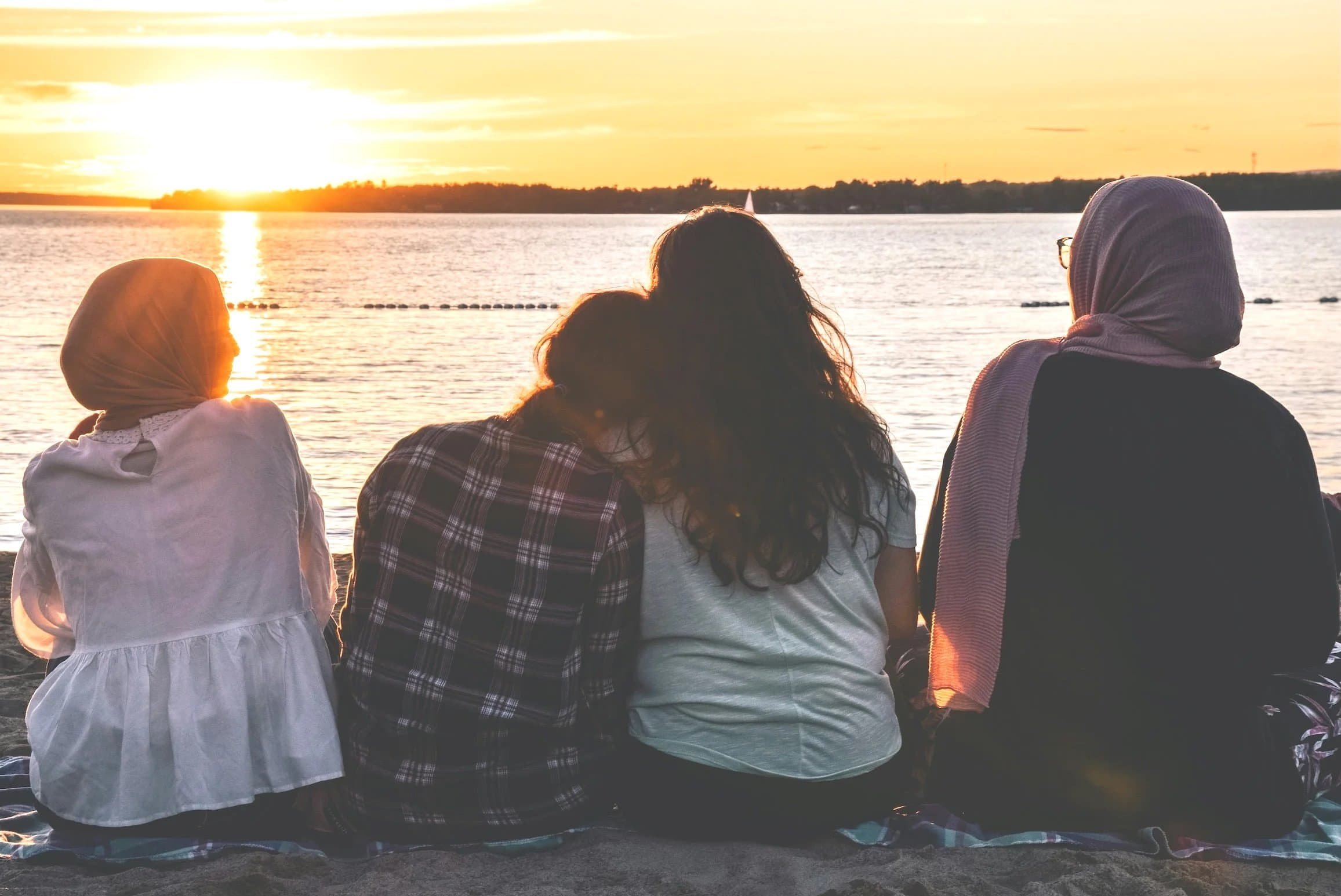 Four people sit on the beach facing a lake at sunset, with their backs to the camera.