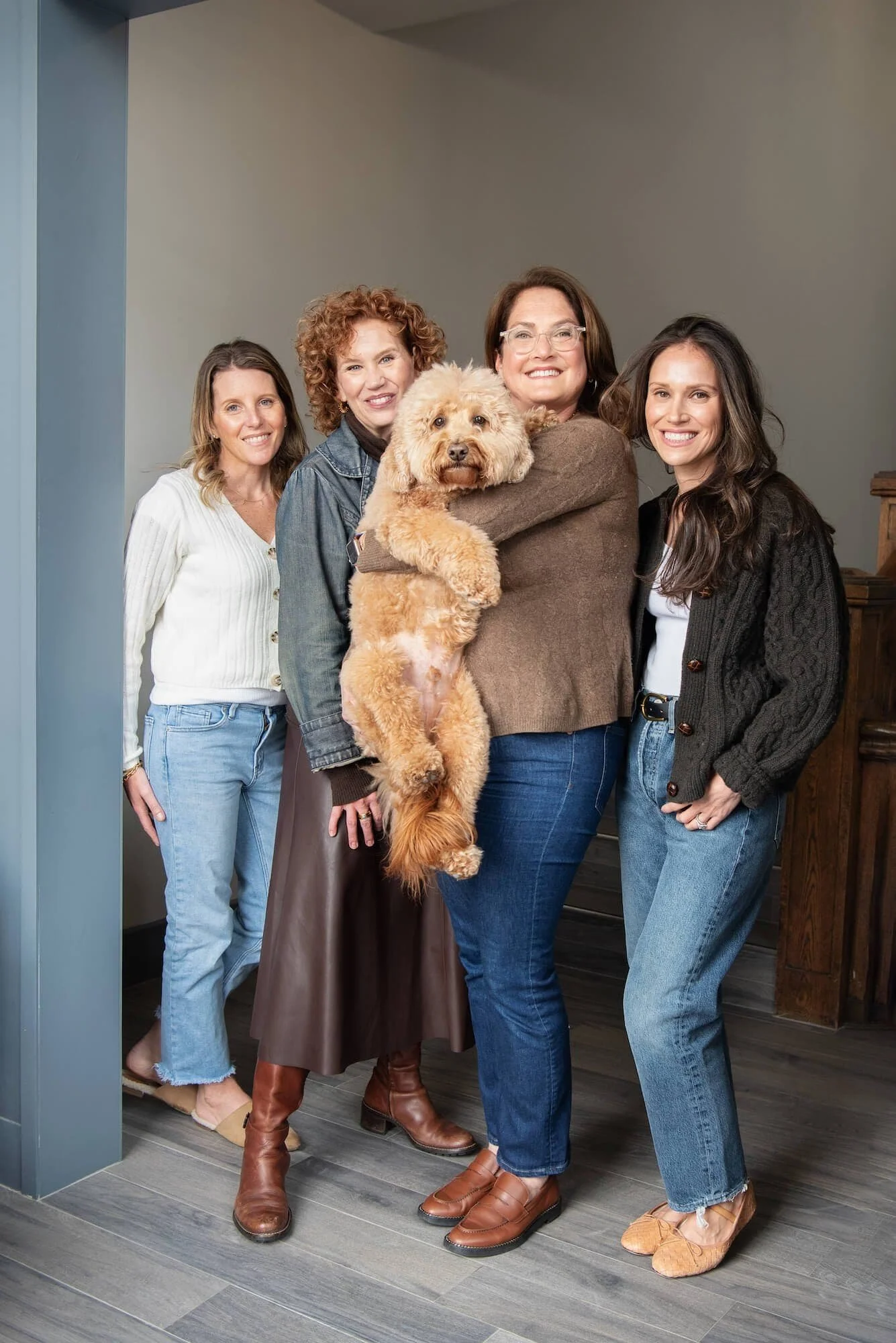 Four women smile at the camera indoors; one woman in the center is holding a fluffy tan dog.