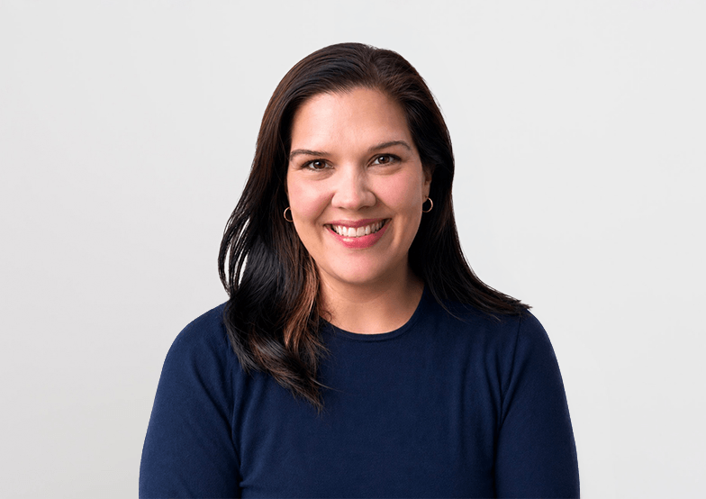 A woman with dark shoulder-length hair, smiling, wearing a navy blue shirt, standing against a plain white background.