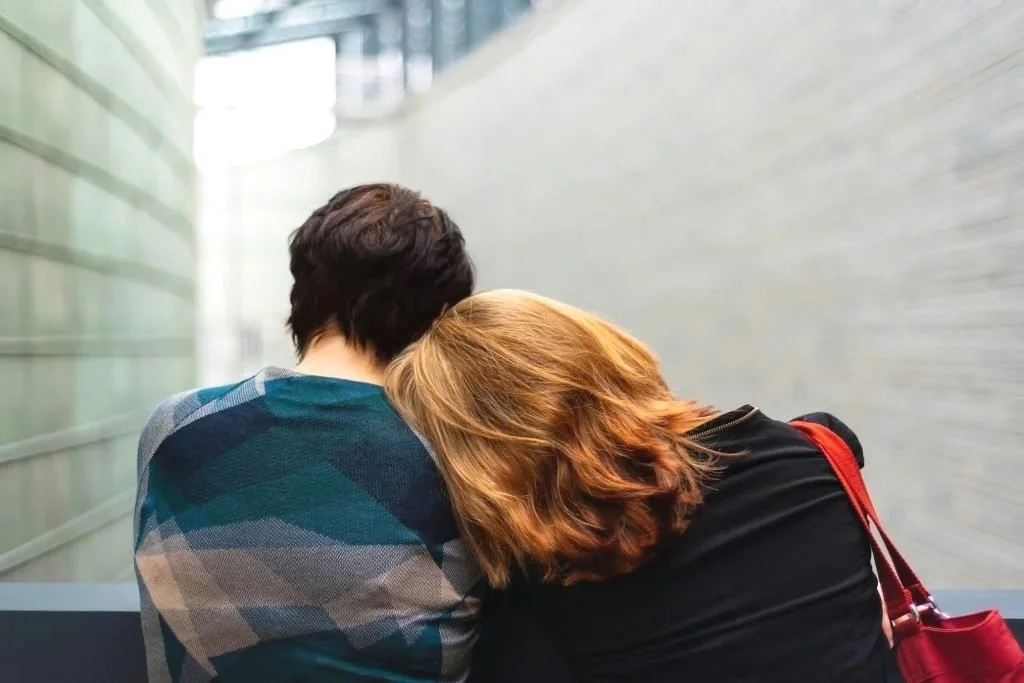Two people sit close together with one resting their head on the others shoulder, seen from behind in a modern hallway.