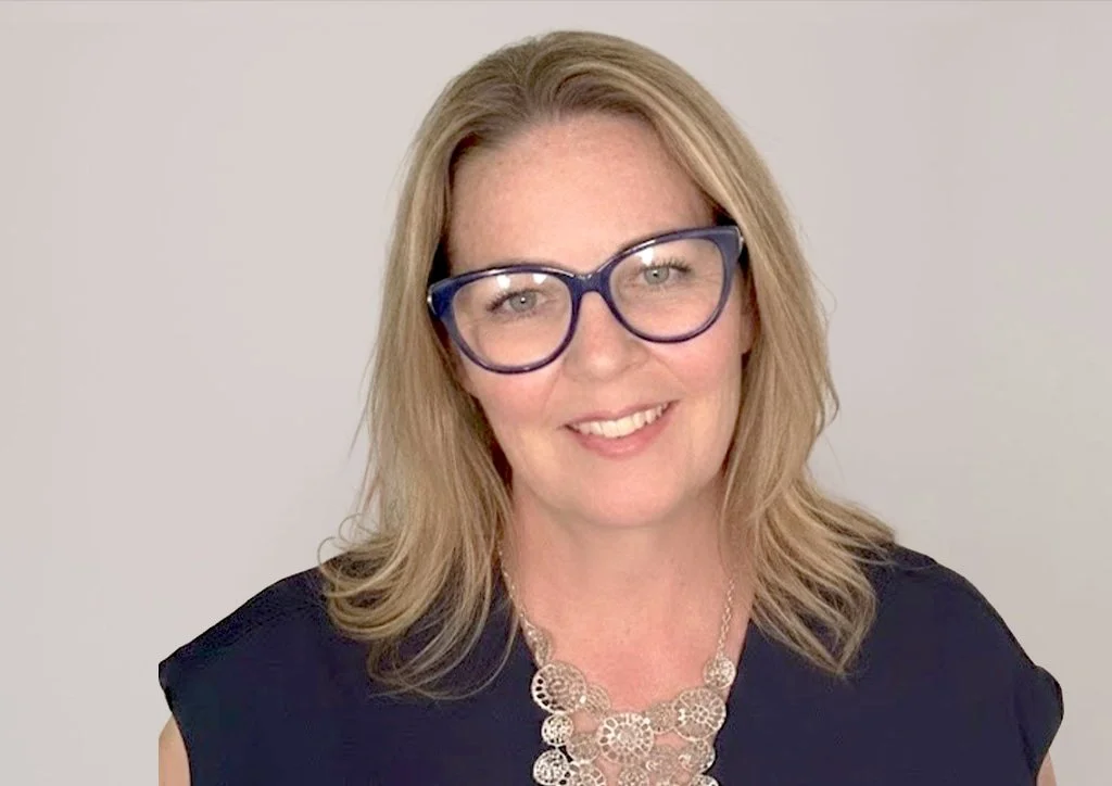 A woman with blonde hair wearing glasses and a black top, smiling against a plain light background.