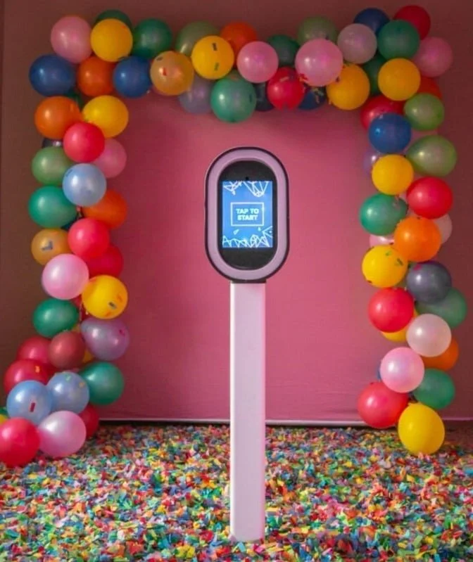 A colorful display with a pink wall and a floor covered in multicolored confetti. In front, there is a hand sanitizing station with a screen saying 'Tap to Start,' surrounded by an arch of multicolored balloons.