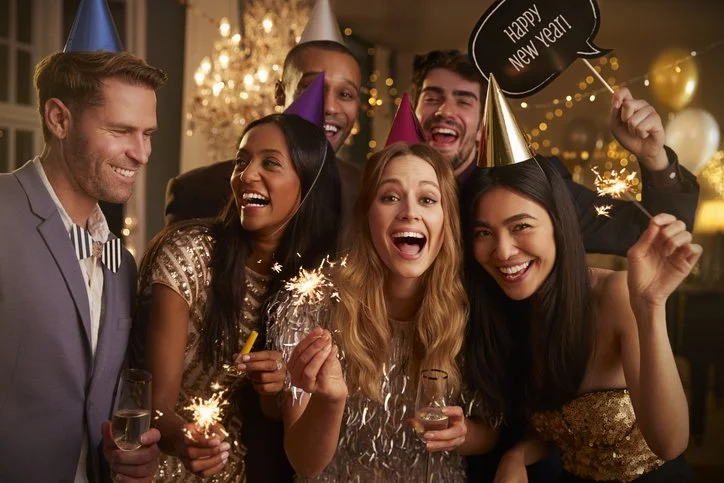 Group of six friends celebrating New Year's Eve, wearing party hats and holding sparklers and drinks, with a festive background.