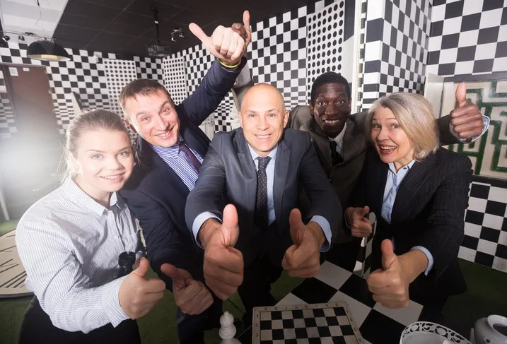 Group of five people in business attire smiling and giving thumbs up inside a black and white checkered room, resembling a chessboard pattern.