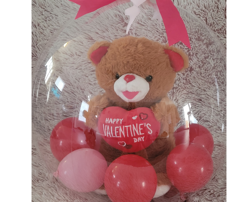 Teddy bear holding a heart-shaped balloon with 'Happy Valentine's Day' written on it, surrounded by pink and red balloons, inside a clear plastic display with a pink ribbon. The teddy bear is placed on a soft, shaggy surface.