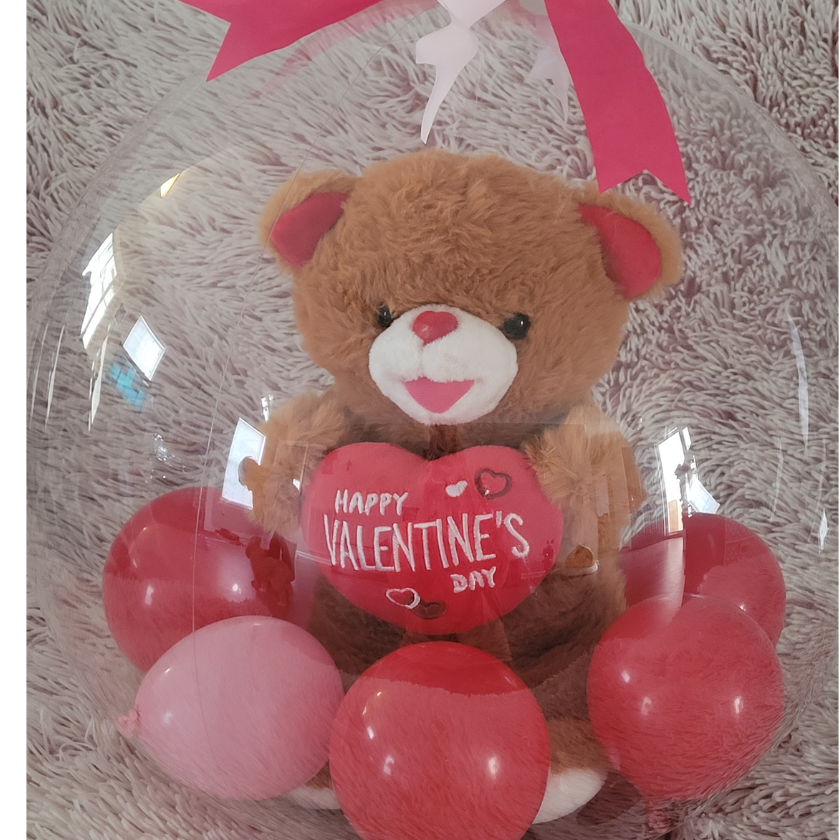 Teddy bear holding a heart-shaped balloon with 'Happy Valentine's Day' written on it, surrounded by pink and red balloons, inside a clear plastic display with a pink ribbon. The teddy bear is placed on a soft, shaggy surface.