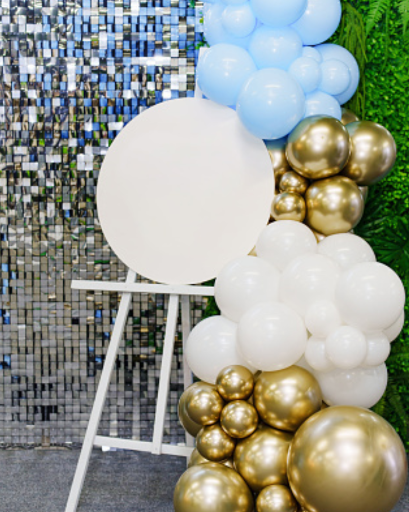 Decorative balloon arrangement with white, gold, and light blue balloons in front of a mirror and green foliage backdrop.