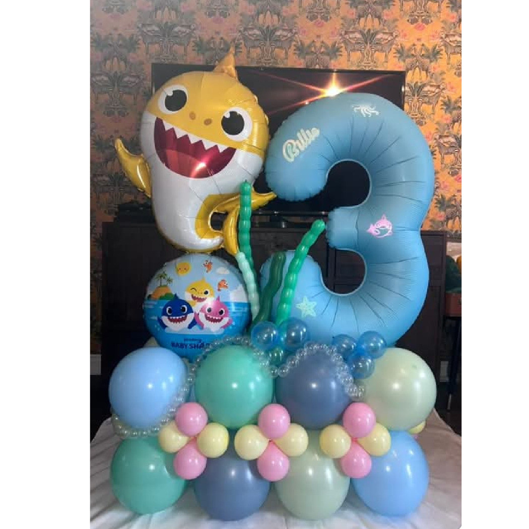 Colorful ocean-themed balloon display with a large blue number 3, a yellow shark character balloon, and a smaller underwater scene balloon showing two sharks. The display features various pastel-colored balloons arranged to resemble an underwater sce