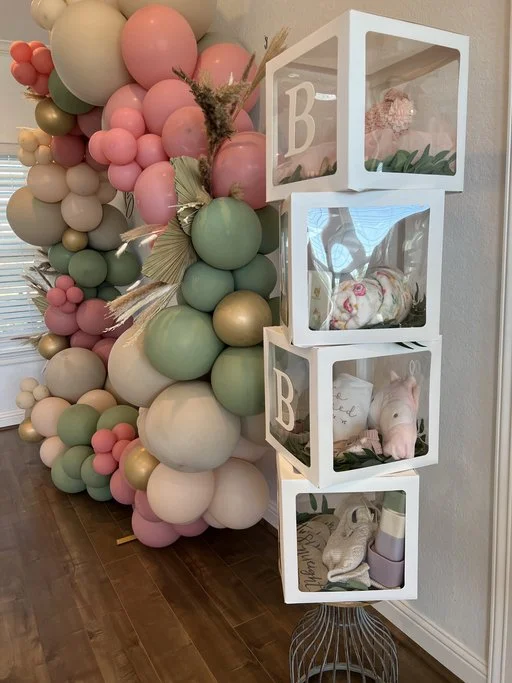 Decorative display with pastel balloons in pink, green, cream, and gold, alongside white cube shelves containing wrapped gifts and plush toys, set against a white wall and wooden floor.