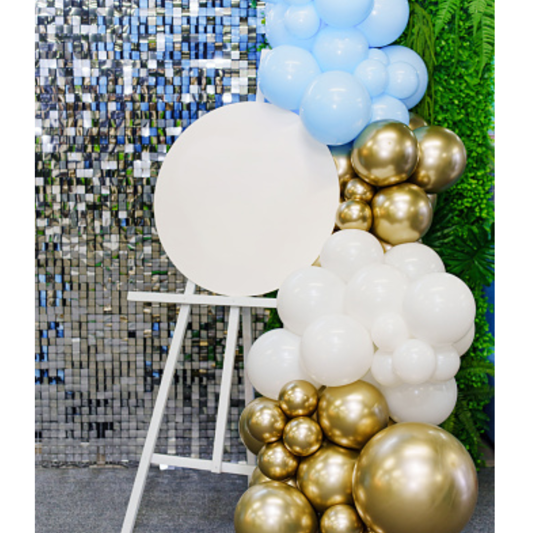 Decorative balloon arrangement with white, gold, and light blue balloons in front of a mirror and green foliage backdrop.