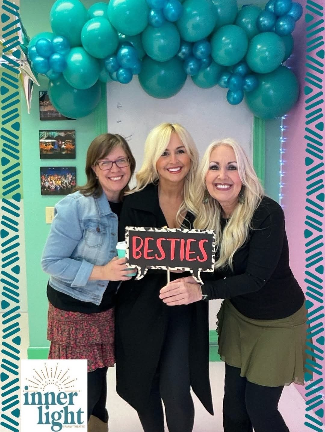 Three women standing together smiling at a celebration, holding a sign that says 'Besties', with a turquoise balloon arch overhead and framed photos on the wall behind them.