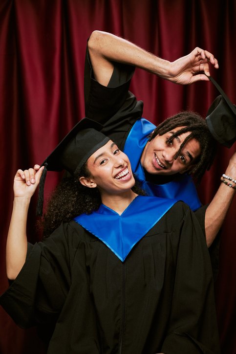 Two graduates in caps and gowns celebrating together, smiling, with a red curtain background.