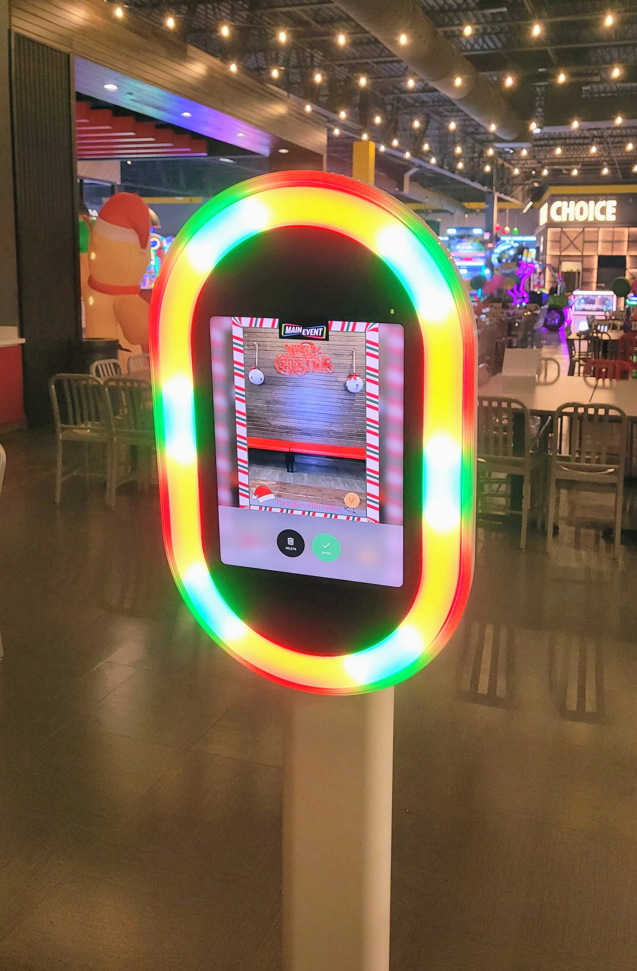 A digital photo booth with a rainbow-colored LED border inside an indoor arcade or entertainment center. The booth has a touchscreen displaying a photo of a decorated background with candy cane borders, a small gingerbread cookie icon, and buttons for deleting or saving the photo. The background features arcade games and a seating area with chairs and tables, illuminated by string lights.