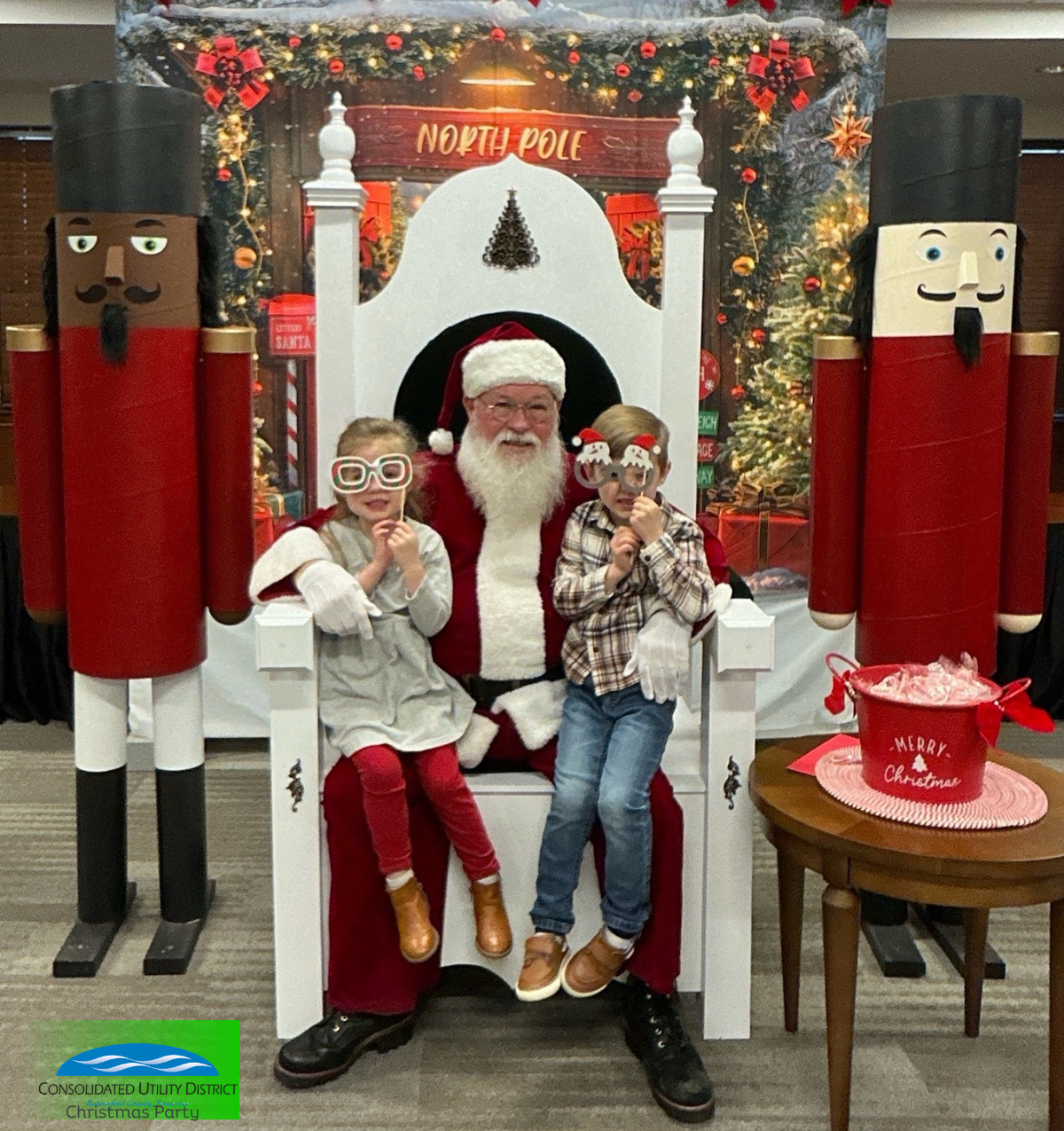 Children sitting on Santa's lap during a Christmas party at the North Pole themed backdrop with nutcracker decorations, Christmas tree, and a "Merry Christmas" bucket, with a man dressed as Santa Claus.