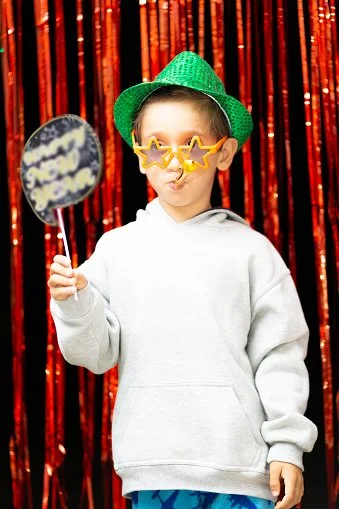 A young boy wearing a green hat and star-shaped sunglasses holding a speech bubble sign, standing in front of a red sequin curtain.