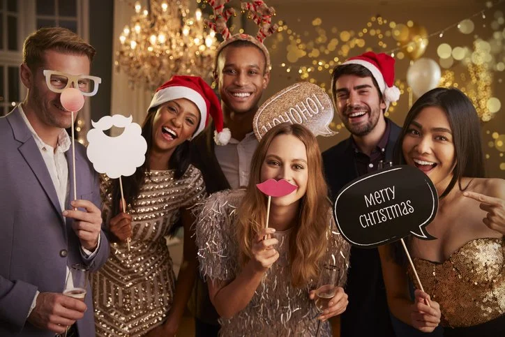 Group of six friends celebrating Christmas, wearing festive hats and costumes, holding party props like a fluffy beard, sunglasses, speech bubbles with Christmas greetings, and smiling indoors with holiday lights and chandelier in the background.