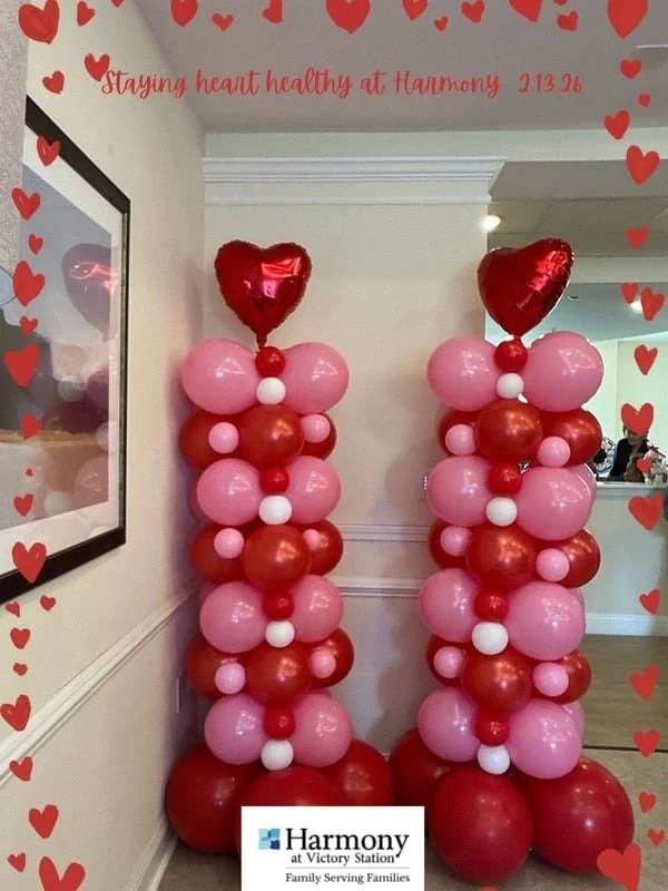 Two balloon columns decorated with pink, red, and white balloons, topped with red heart-shaped balloons, inside a room with a wall and a picture frame, with a border of red hearts and a quote about staying heart healthy.