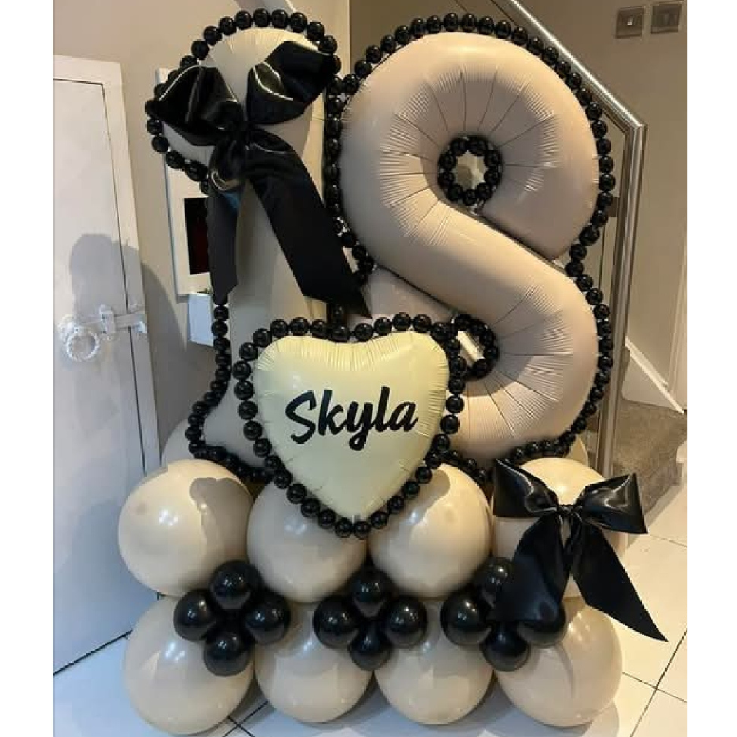 Balloon arrangement featuring a large beige number "5", a heart-shaped balloon with the name "Skyla" written on it, black and beige balloons, and black ribbon bows.