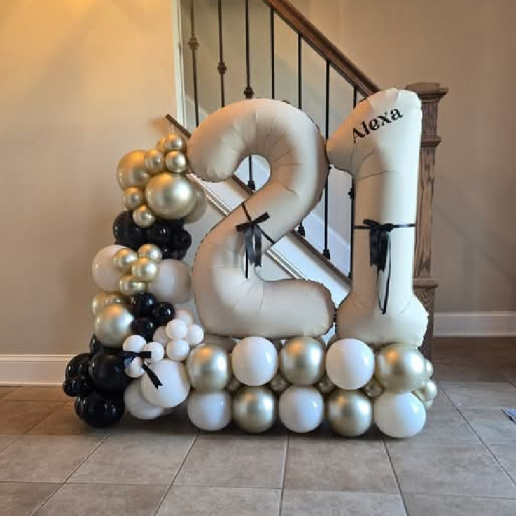 Decorative balloon sculpture of the numbers '21' with black, white, and gold balloons, placed on the floor in front of a staircase. The number '21' is made of large balloons, and the sculpture includes smaller balloons clustered at the base and on th