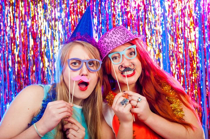 Two women wearing colorful costumes and funny accessories like glasses, foam mustaches, and party hats at a party with bright, shiny, multicolored streamers in the background.