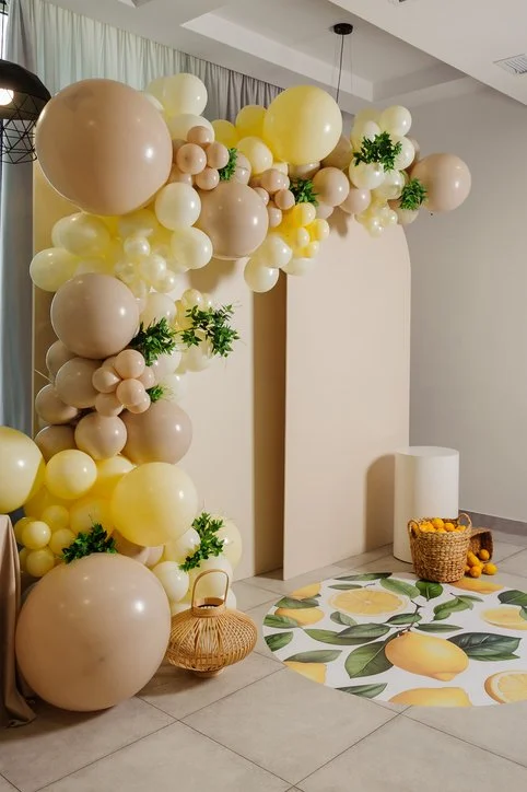 Indoor decorative balloon arrangement with beige, cream, and yellow balloons, some with greenery, in front of a beige wall with a white and lemon-themed rug on the floor and a basket of lemons nearby.