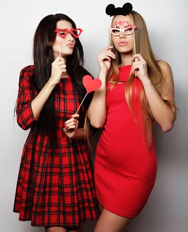Two women standing together holding playful photo booth props, one with red heart-shaped glasses and a heart stick, the other with pink 'Mrs.' glasses and a pig nose stick, dressed in red and plaid outfits.