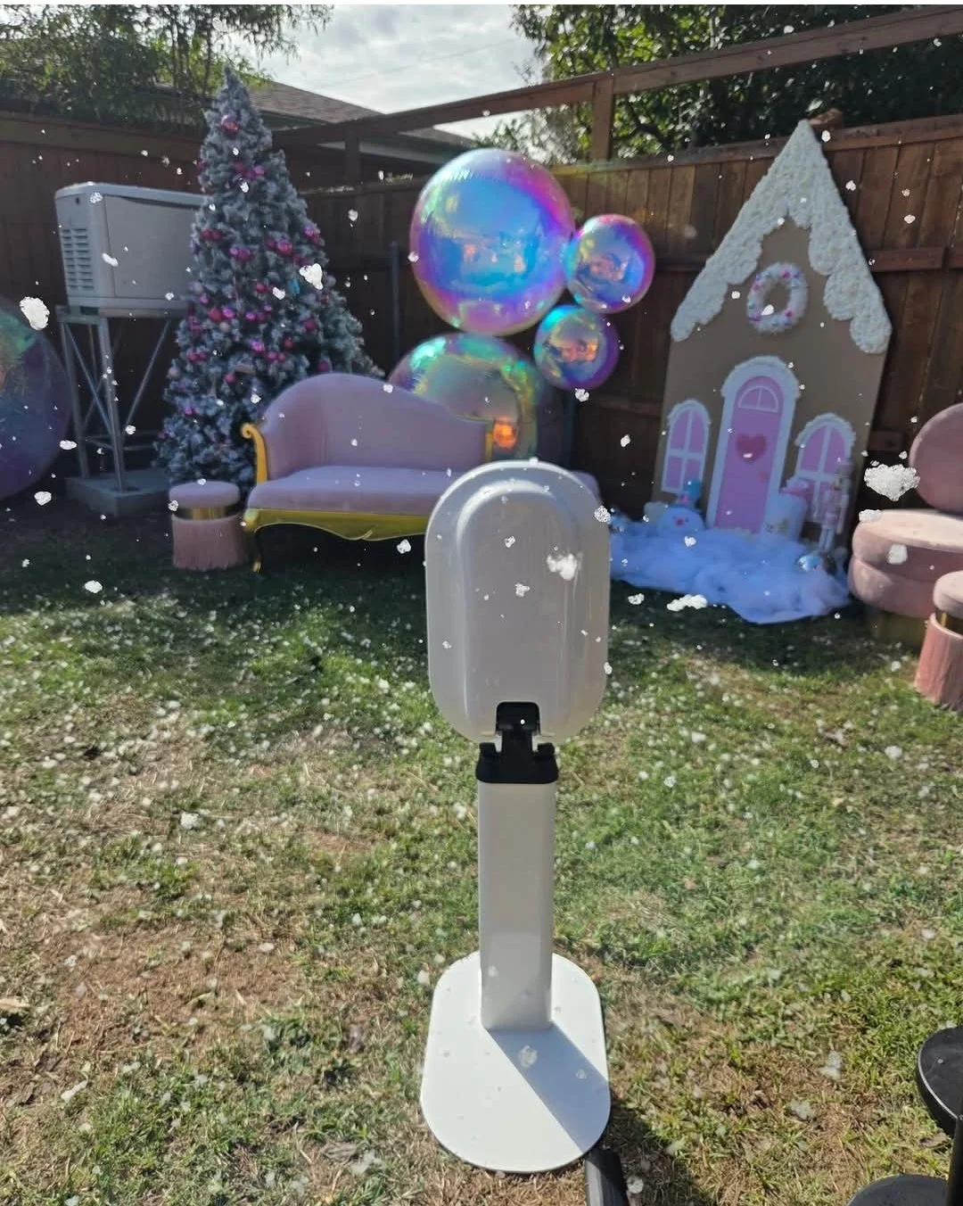 A backyard scene with Christmas decorations, including a Christmas tree, colorful balloons, and a small house-shaped decoration, with soap bubbles floating in the air and a white stand in the foreground.