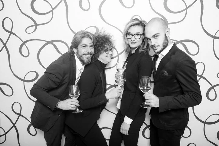 Four people at a party holding wine glasses, standing against a patterned wall, dressed in formal attire, smiling and posing for the camera.