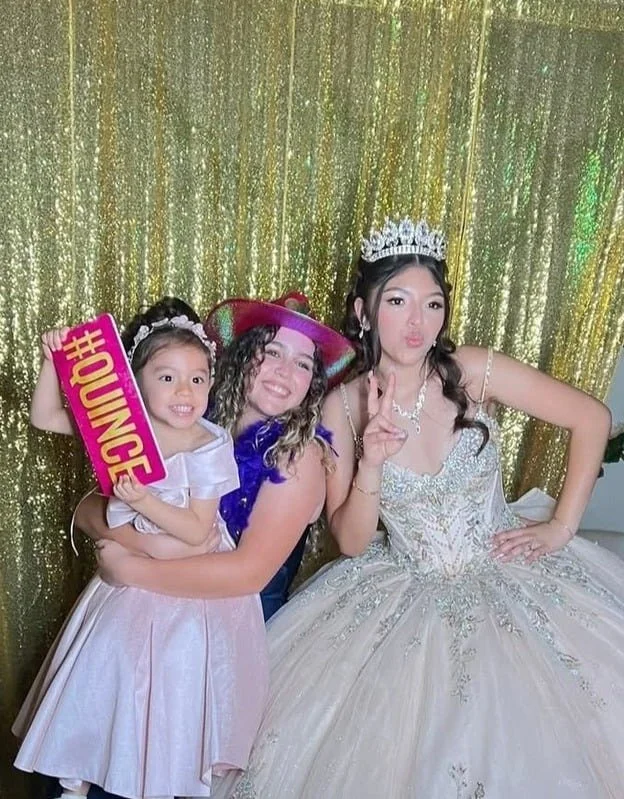 Three women at a celebration, with a girl holding a pink sign that says '#HUNGE'. The woman on the right wearing a tiara and white dress, the woman in the middle with a colorful hat and purple dress, and the girl in a light pink dress.