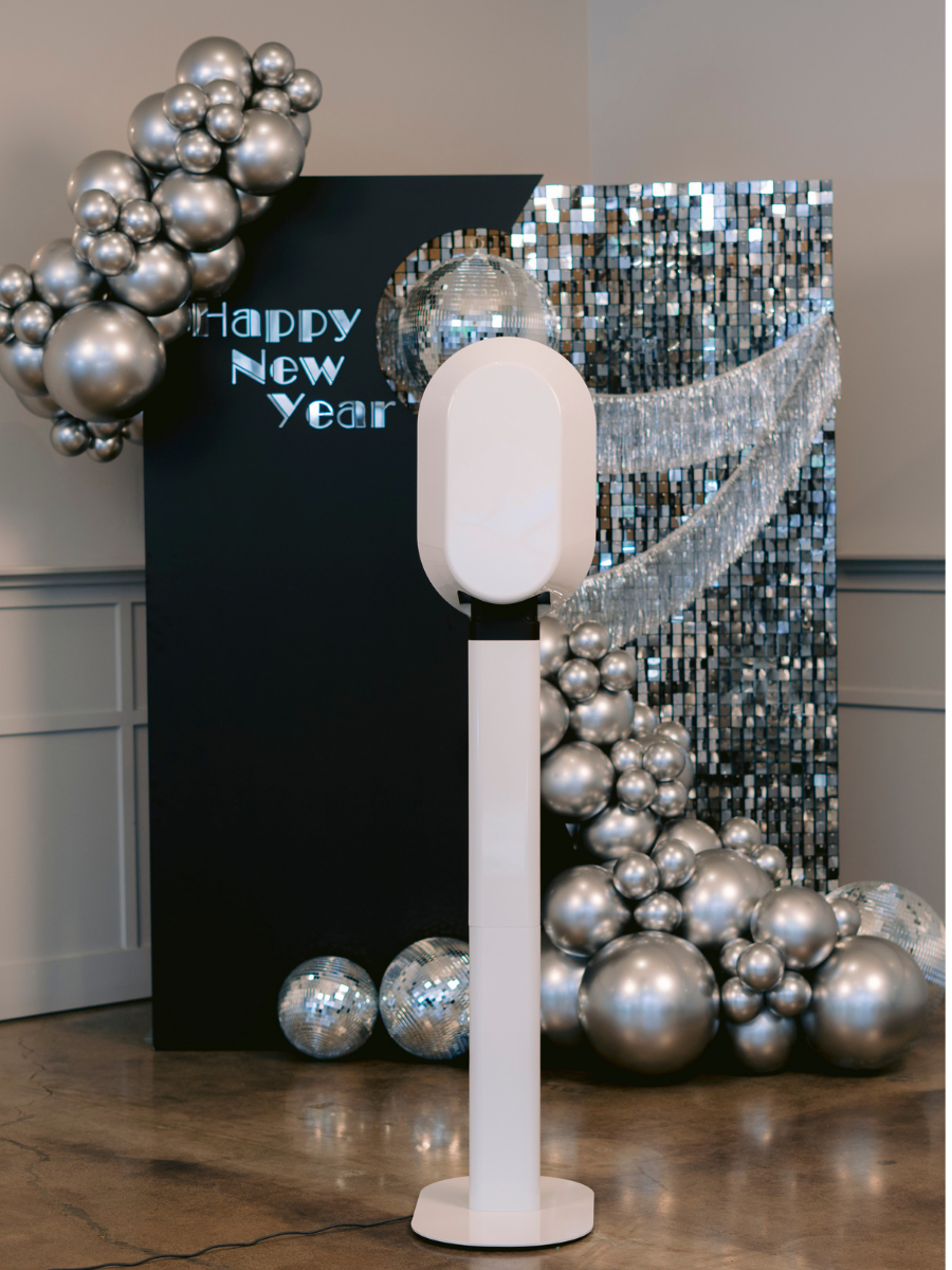A New Year's celebration display with silver balloons, a dark backdrop with bright text saying 'Happy New Year,' a disco ball, and a large white air purifier or fan in front.