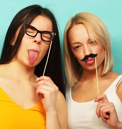 Two women holding celebrity-style props: one with glasses and a stick, sticking out her tongue, and the other with a fake mustache on a stick, making a playful face.
