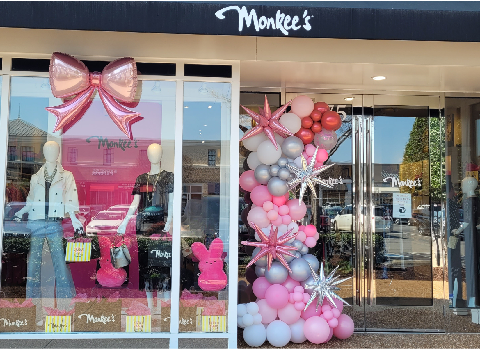 Storefront with Monkee's logo, window display with mannequins dressed in clothing, pink and silver balloon decorations including bows, stars, and bunnies outside the entrance.