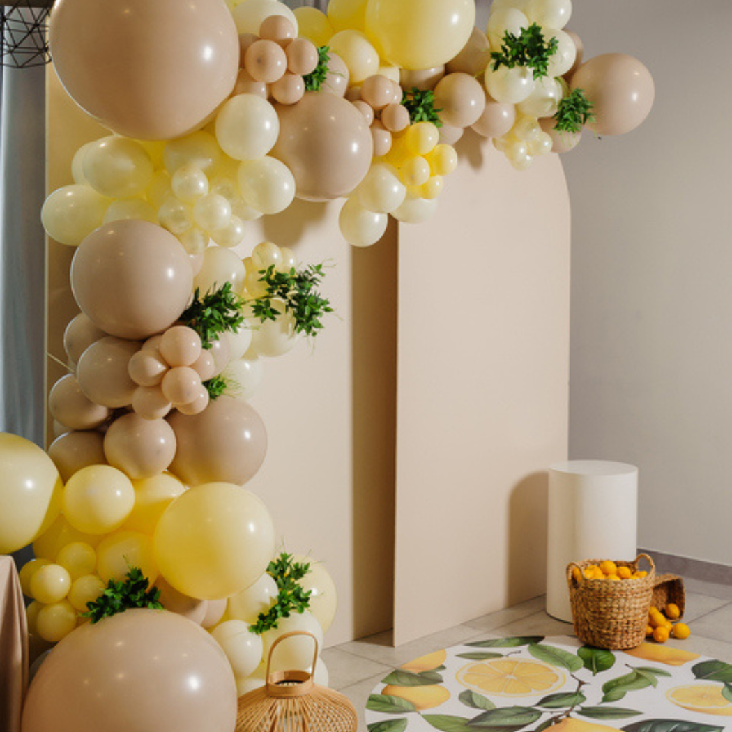 Decorative pastel-colored balloon arch with balloons in shades of cream, beige, and yellow, accented with small green leaves, decorating a wall near a round table with lemon print, set in a room with neutral walls and flooring, featuring a wicker bas