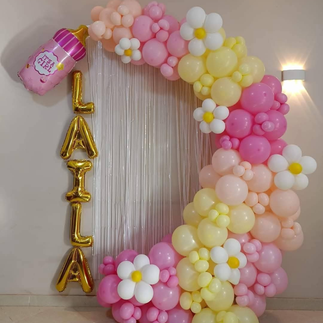 Balloon arrangement with pink, yellow, and white balloons, decorated with white flower balloons with yellow centers, and gold letter balloons spelling 'LAEL'. A pink balloon shaped like a milk bottle with the phrase 'It's a Girl' is attached to the t