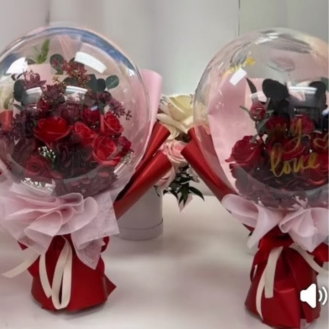 Two bouquets of red roses inside transparent balloons, decorated with pink and red ribbons and wrapping paper, with a background of mixed flowers.