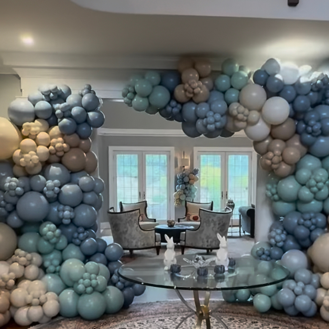 Balloon arch with shades of blue, white, and beige frames a living room area with four chairs, a round table, and a bouquet of balloons in the background.
