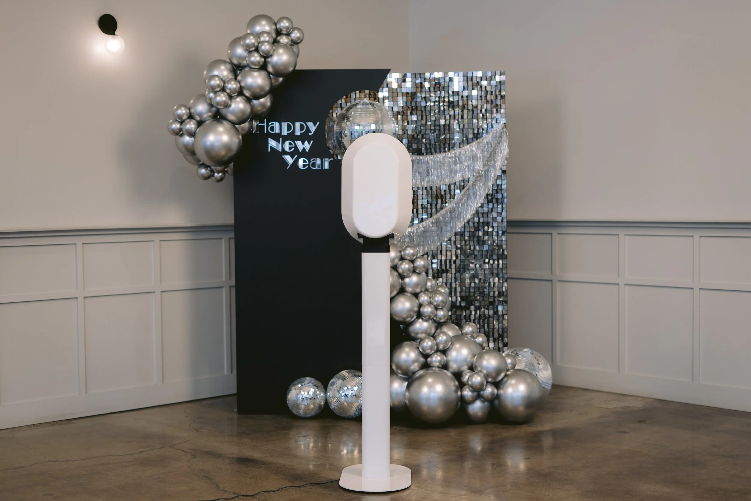 Decorations for New Year's celebration including silver balloons, mirrored disco balls, a black panel with a 'Happy New Year' sign, and a white photo booth stand, all set against a wall with wainscoting.