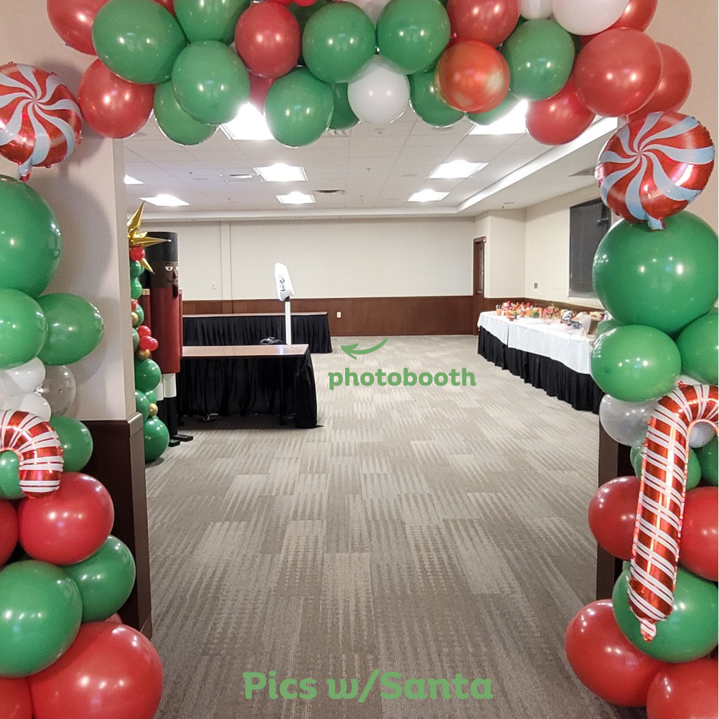 Decorated entrance with arch of red, green, white, and striped balloons at a holiday party. Photo booth area in the background with table of treats.